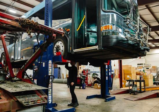 Bus being lifted in a repair shop by hydraulic jacks, with a mechanic underneath.