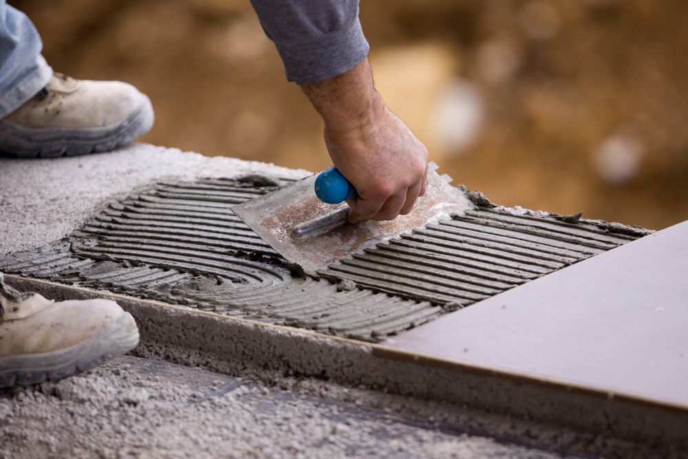 A Man Is Laying Tiles On The Floor With A Trowel — DCK Concreting In Port Macquarie, NSW