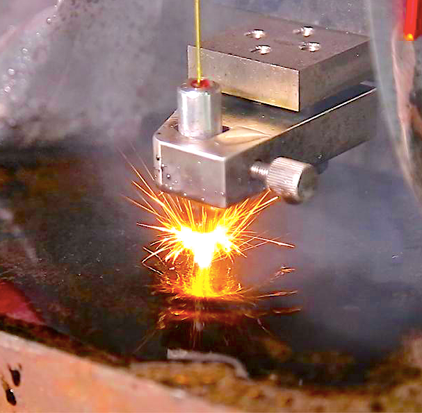 Una boquilla industrial metálica corta una placa oscura, produciendo chispas de color naranja brillante y material fundido.