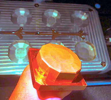 Una mano sostiene una gema de resina facetada de color ámbar frente a un molde de silicona multicavidad a juego.