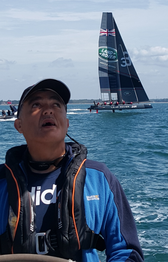 A close up picture of a VI male sailor at the wheel of an underway yacht. In the background is a racing yacht of the Land Rover BAR team, skippered by Ben Ainslie, passing at high speed raised on aerofoils. A close up picture of a VI male sailor at the wheel of an underway yacht. In the background is a racing yacht of the Land Rover BAR team, skippered by Ben Ainslie, passing at high speed raised on aerofoils.