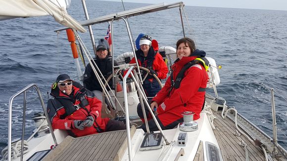 A picture taken from the fore deck of an underway yacht, looking back to the stern. A mixed female and male VI and sighted crew, with a female VI sailor at the wheel, are looking towards the camera, all smiling. A picture taken from the fore deck of an underway yacht, looking back to the stern. A mixed female and male V.I. and sighted crew, with a female V.I. sailor at the wheel, are looking towards the camera, all smiling.