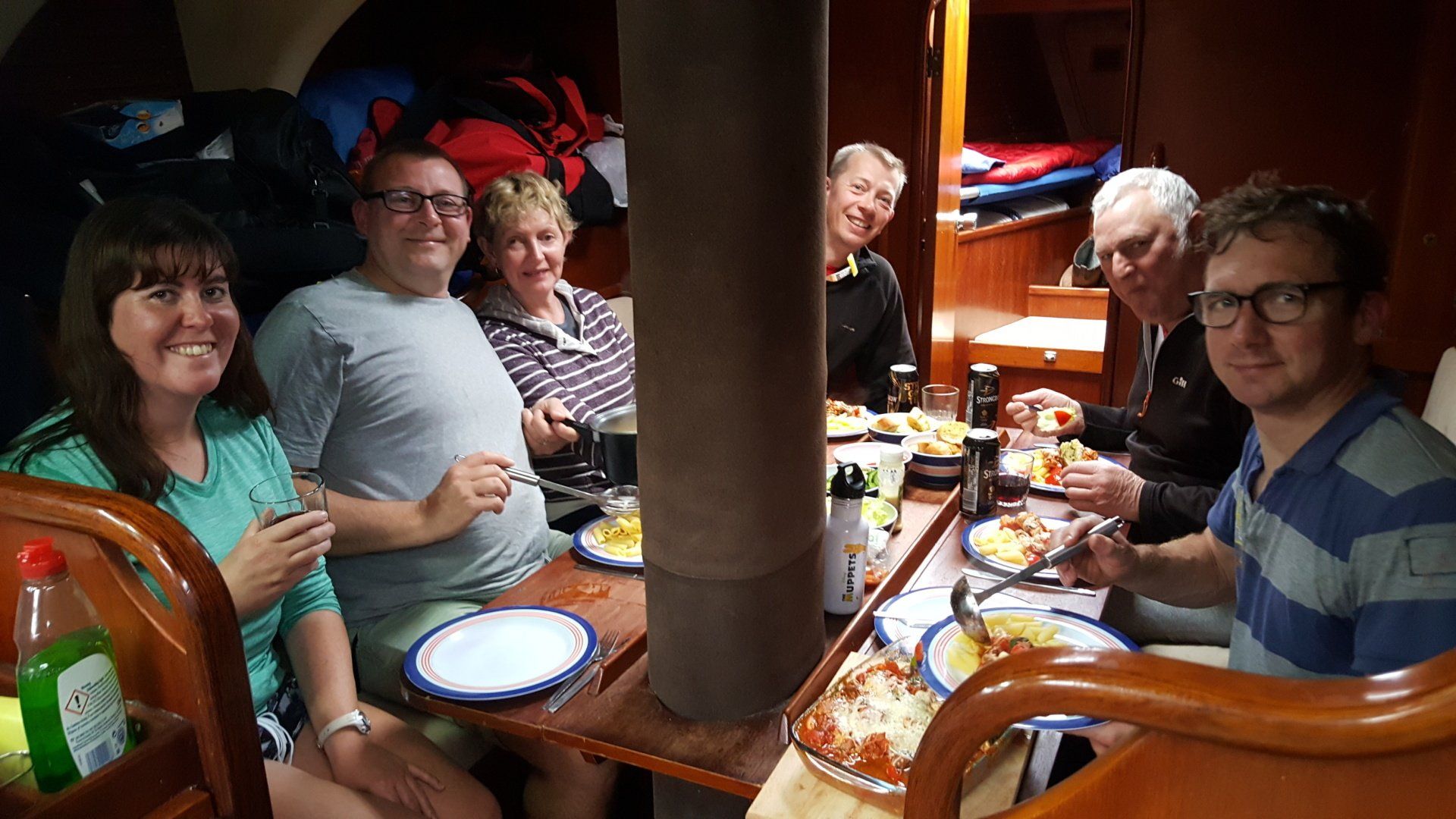 A picture below decks of a crew having dinner. The table is laid with various dishes andd drinks, and everyone is smiling towards the camera.