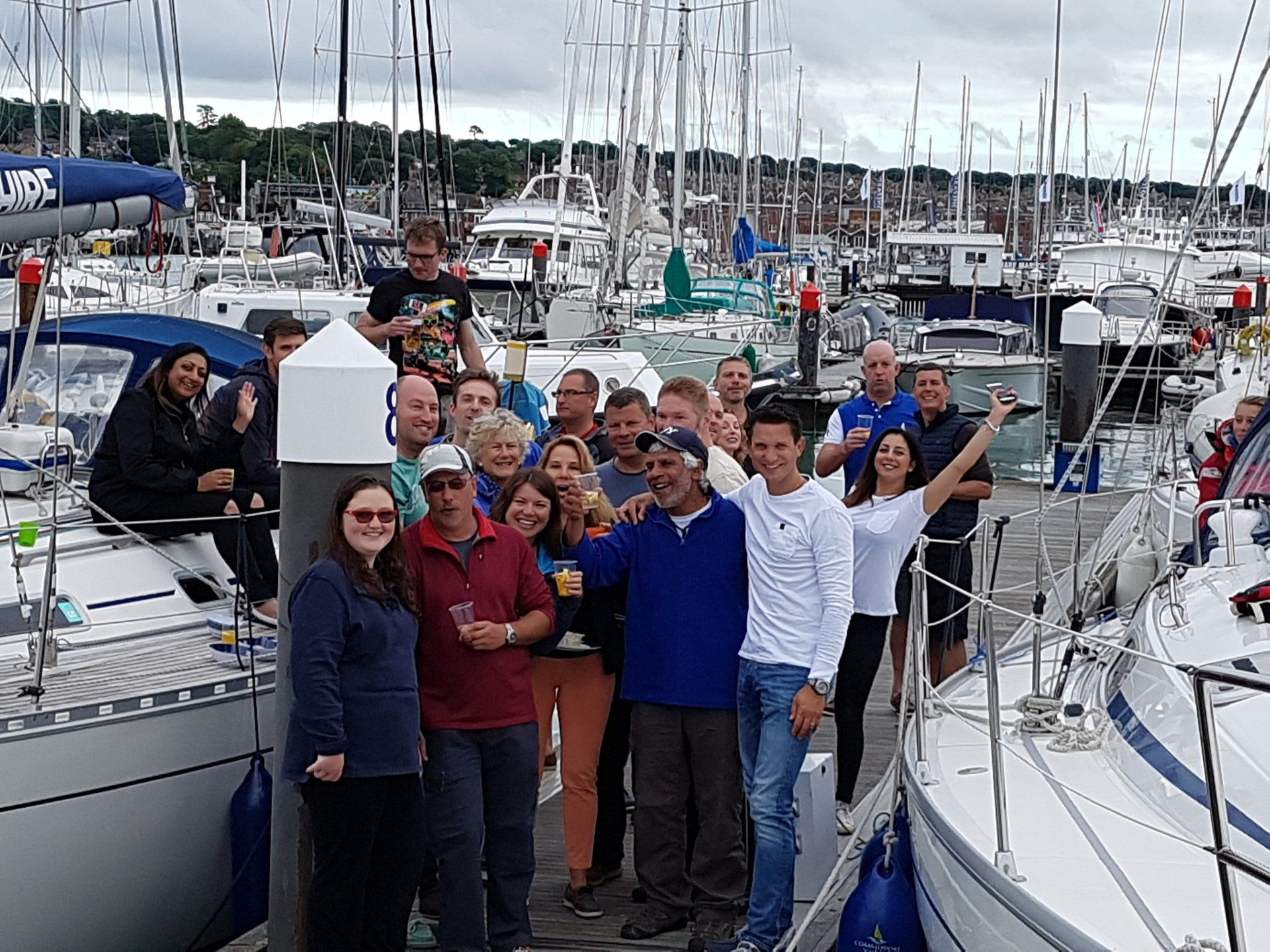 An end-of-day picture of several crews getting together at the marina. Around twenty V.I. and sighted sailors are standing on a pontoon or the deck of the moored yacht, smiling and raising glasses towards the camera.