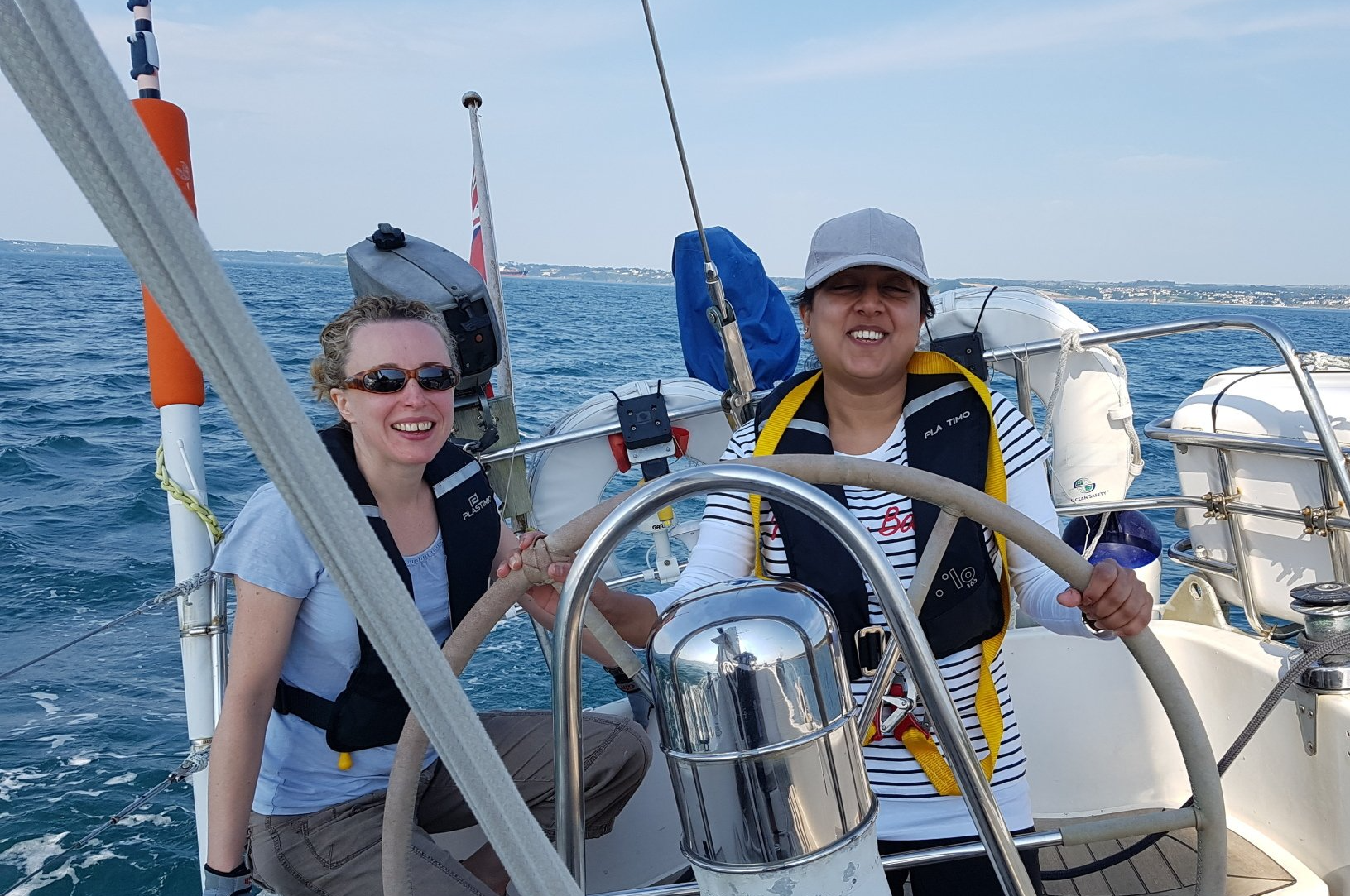A picture of a smiling female VI sailor at the wheel, and a smiling female sighted crew member next to her. Calm conditions. A picture of a smiling female VI sailor at the wheel, and a smiling female sighted crew member next to her. Calm conditions.