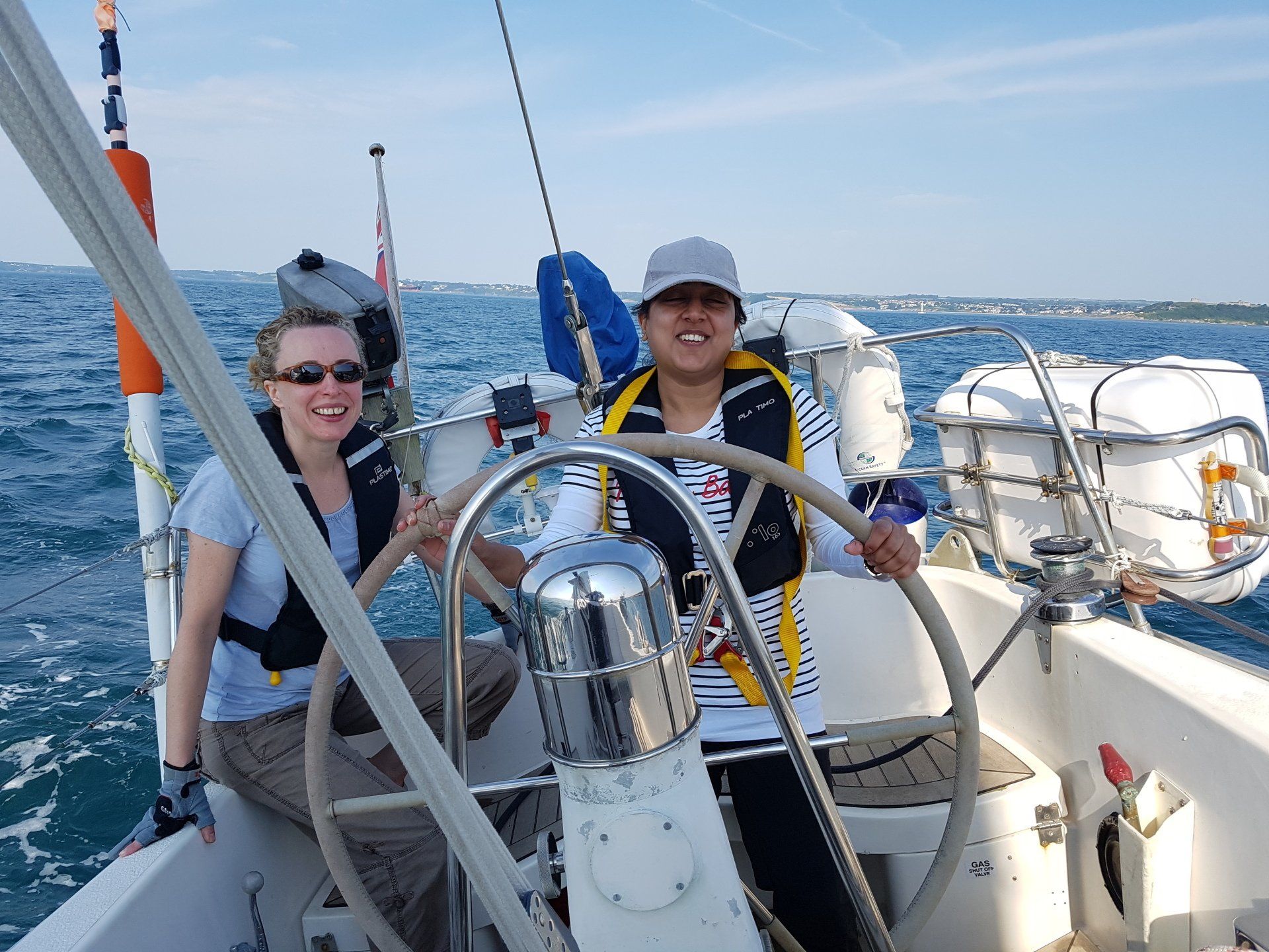 A close up taken from the deck of an underway yacht, looking back to the wheel. The weather is clear. The yacht is being helmed by a V.I. sailor, with a sighted crew member sitting next to them.
