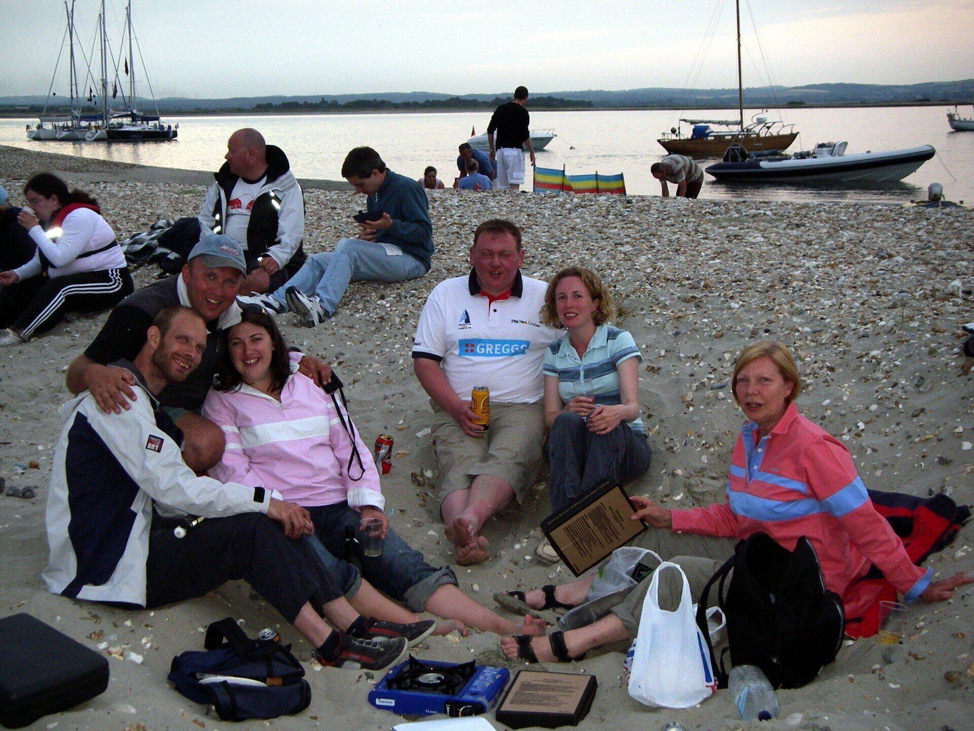A picture of a number of sighted and V.I. sailors relaxing on a shingle beach in the evening. The sea and some moored yachts are in the background.