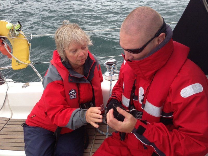 A close up picture taken on board a yacht, of a sighted crew member assisting a V.I. sailor with some wet weather gear and gloves.
