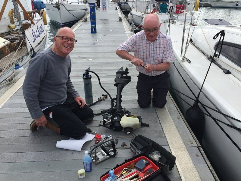 A picture of two sailors kneeling down on a marina pontoon with. yacht alongside. They are working on a small outboard engine, surrounded by tools and equipment. They are looking towards the camera and laughing.