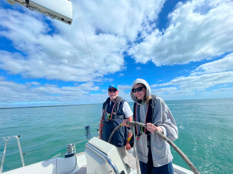 A picture of a sailing yacht underway, taken from the deck looking towards the stern. There is a VI crew member at the wheel with their skipper stood next to them both smiling A picture of a sailing yacht underway, taken from the deck looking towards the stern. There is a VI crew member at the wheel with their skipper stood next to them both smiling