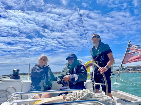 A picture of three crew members, VI and sighted, two sitting on the benches and one stood at the wheel, smiling in front of blue skies.