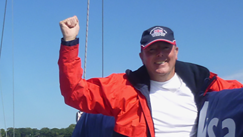 A picture taken from the deck of an underway yacht, looking back to the wheel, being helmed by a visually impaired male sailor. A female sighted crew member sits next to him. The sea and horizon in the background are on an angle, showing the vessel is under sail. A picture taken from the deck of an underway yacht, looking back to the wheel, being helmed by a visually impaired male sailor. A female sighted crew member sits next to him. The sea and horizon in the background are on an angle, showing the vessel is under sail.