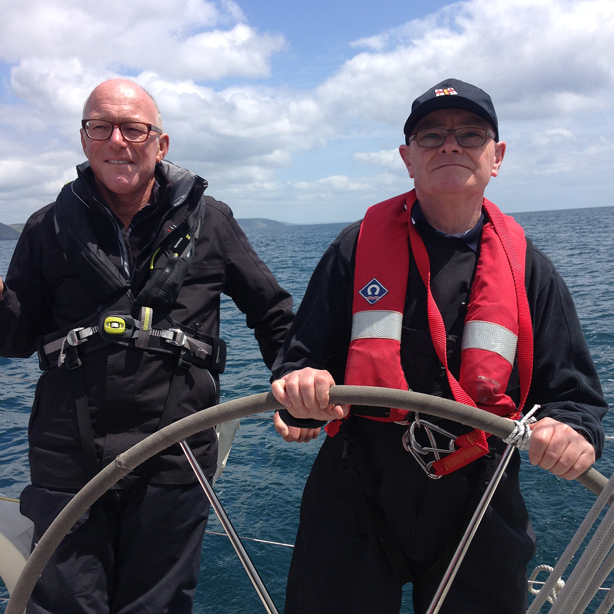 A picture of a sighted male skipper standing next to a male VI sailor who has the wheel. The sea is calm. A picture of a sighted male skipper standing next to a male VI sailor who has the wheel. The sea is calm.