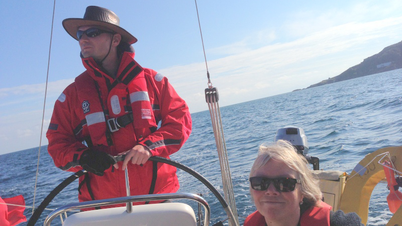 A picture taken from the deck of a yacht, looking back to the wheel. The horizon is at an angle indicating that the yacht is under sail. A V.I. sailor is at the helm, and a smiling sighted crew is sitting next to him.