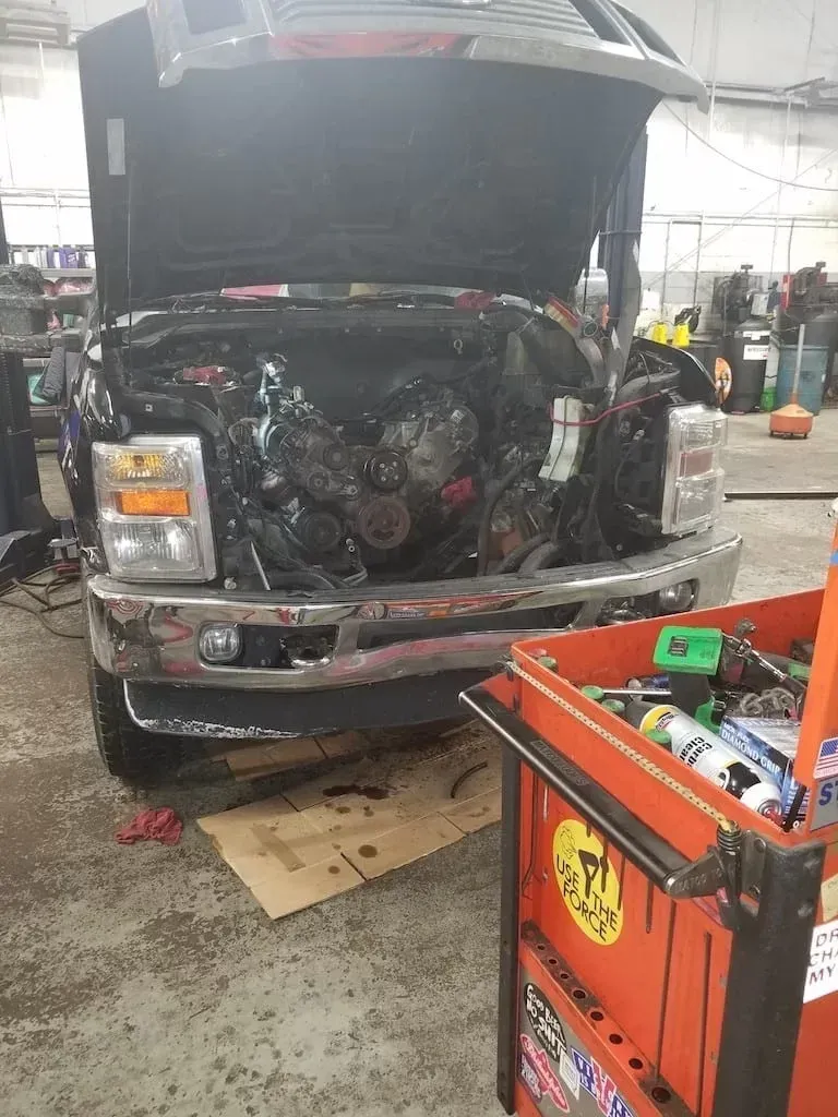 Black truck with hood open in a garage, exposing the engine; orange tool cart in foreground.