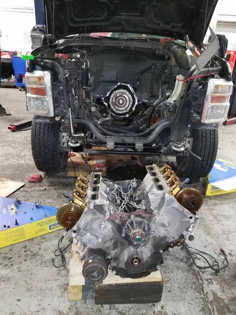 Truck with engine removed in a workshop, engine assembly on the floor in front of it.