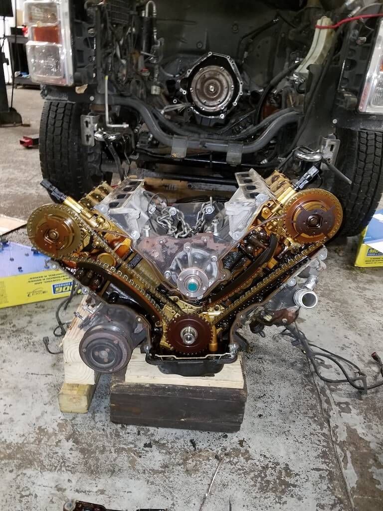 Disassembled car engine with V-shaped polished gold and black timing cover in a garage.