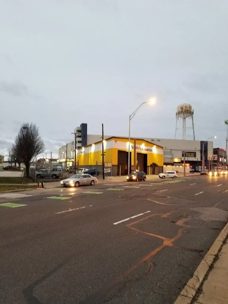 Dusk at a wet street corner with a yellow-lit building, parked cars, and a water tower in the background