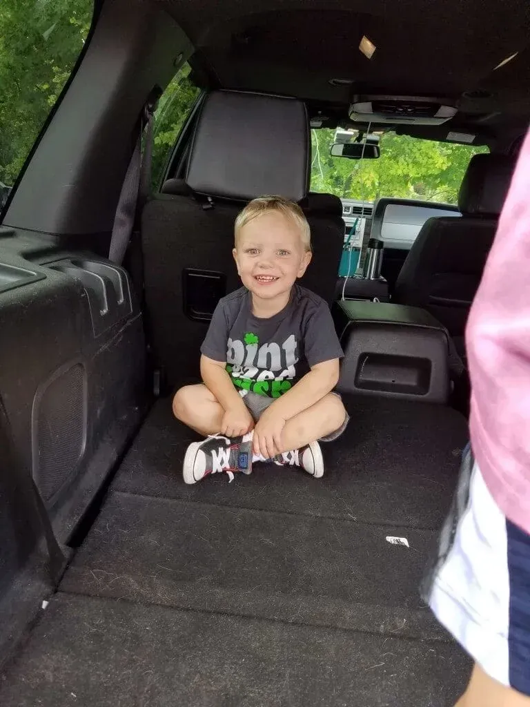 Smiling child sitting cross-legged in the back of a vehicle, wearing a dark shirt and sneakers.
