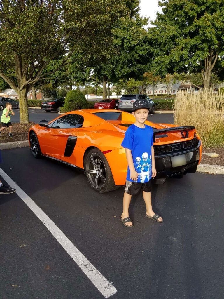 Boy standing in front of an orange sports car in a parking lot, holding a blue toy car-shaped board