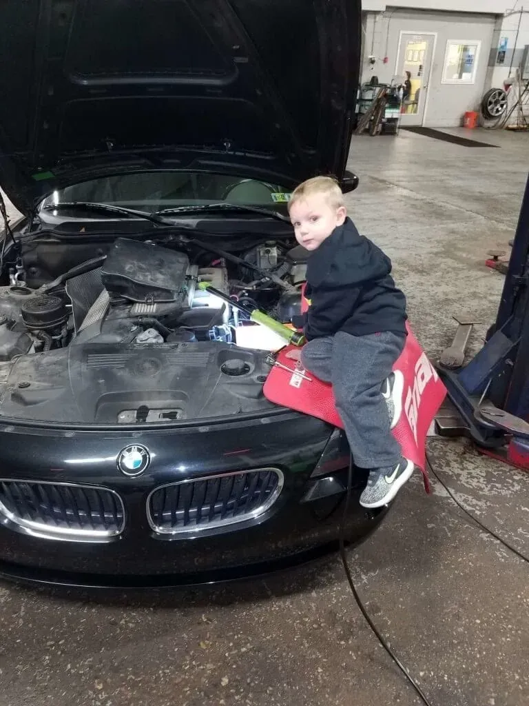 Child sitting on the front of a black BMW in a garage, hood open and engine exposed