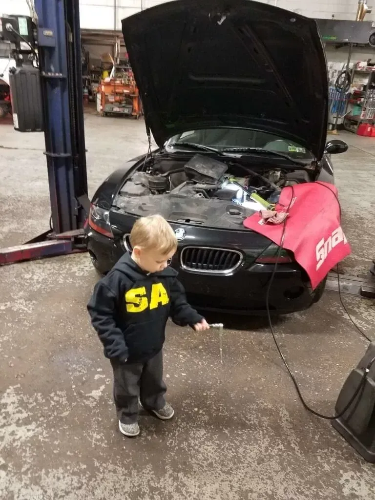 Child standing in an auto shop beside a black BMW with its hood open and a pink fender cover