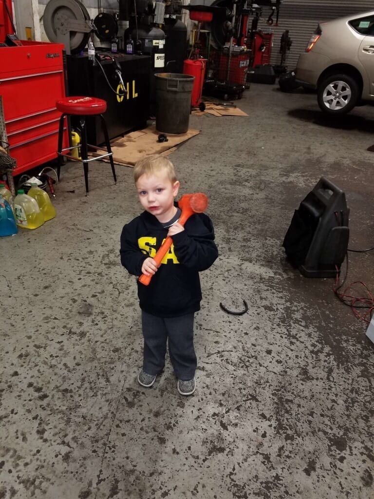 Child in a dark hoodie holding an orange toy tool in a garage, with cars and equipment in the background