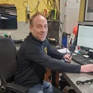 Man sitting at a desk in an office, smiling while holding a phone near a computer and keyboard.