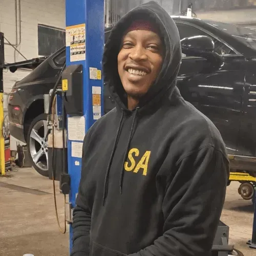 Smiling person in a black hoodie with gold “SA” lettering, standing in an auto shop beside a lifted black car.