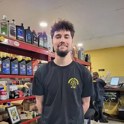 Smiling person in a black shirt standing in an auto parts shop with shelves of oil bottles behind them