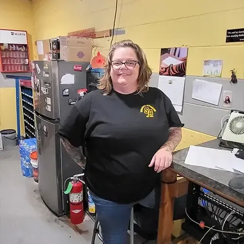 Woman smiling in a workshop, leaning by a workbench with a fire extinguisher and refrigerator nearby.