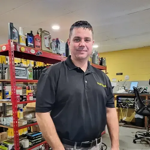 Man in black polo standing in an auto shop beside shelves of oils and tools