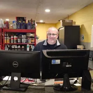 Smiling person behind two computer monitors in a shop, with shelves of products in the background