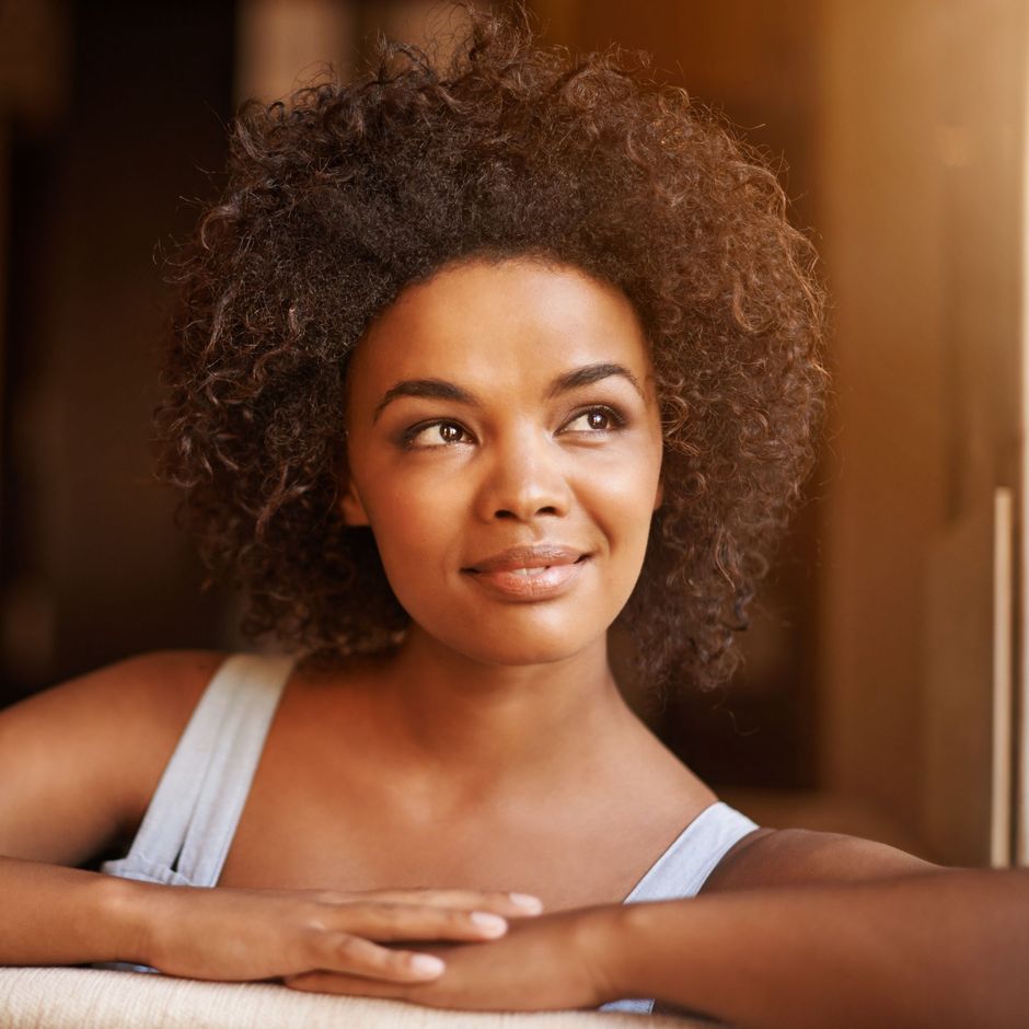 Smiling person with curly hair resting arms on a surface, looking to the side.