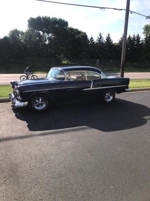 Black 1955 Chevrolet Bel Air parked on asphalt, with a person on a bicycle in the background.