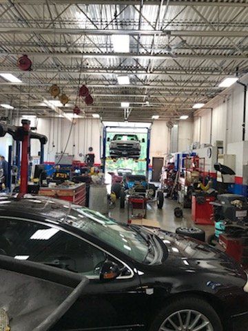 Car repair shop with a vehicle on a lift, mechanic working. Black car in foreground, tools, equipment.
