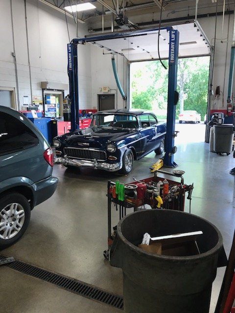 Classic blue car on a lift in a garage with tools and a van, doorway in the background.