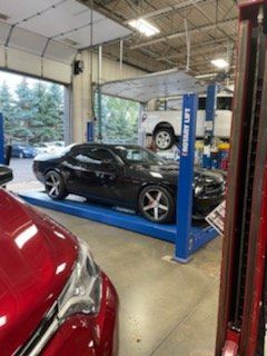 Black Dodge Challenger on a blue car lift inside a car repair shop. A red car is in the foreground.