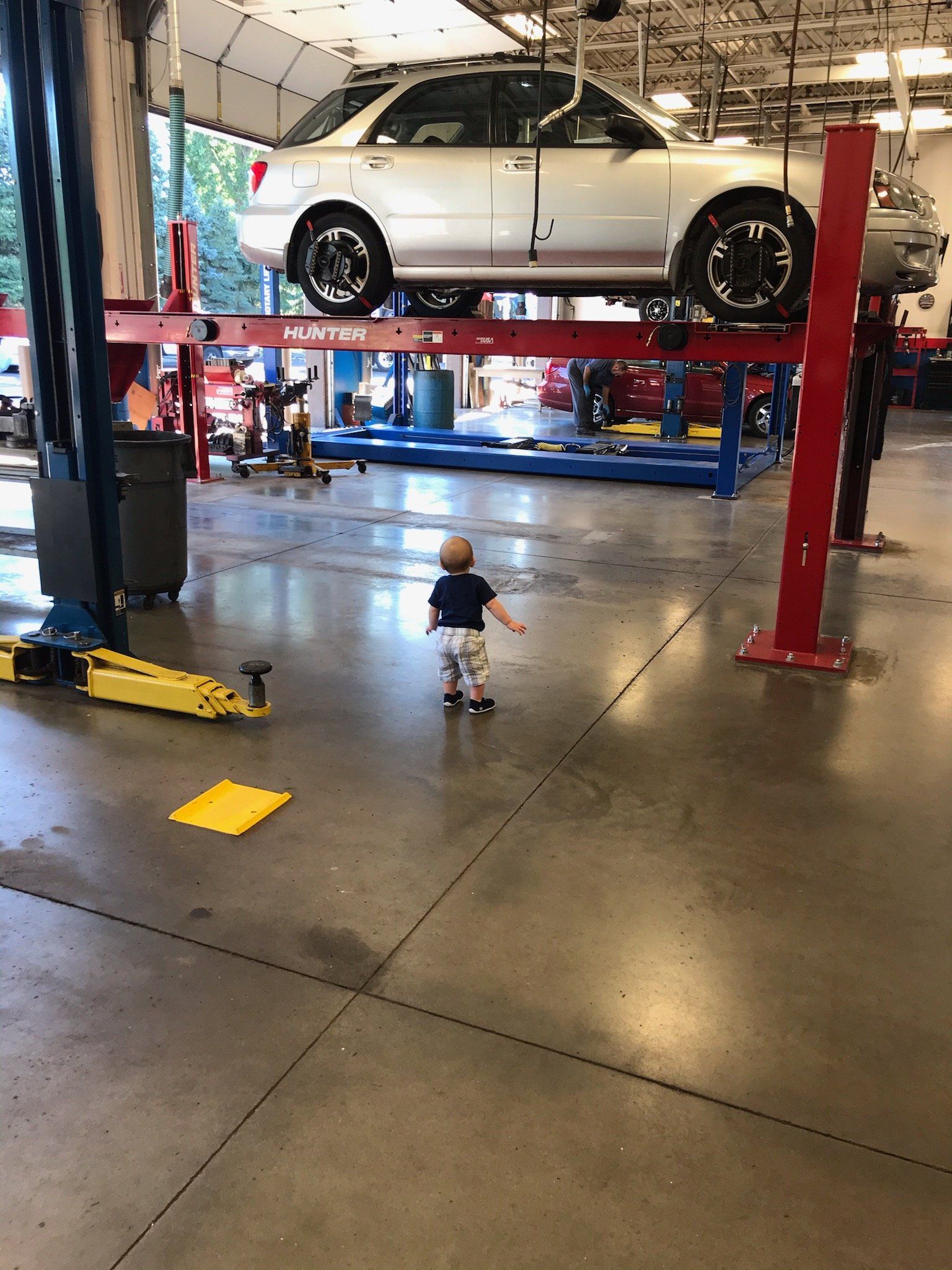 Child walking in a garage with a car lifted in the air by a red lift. Mechanics work in the background.