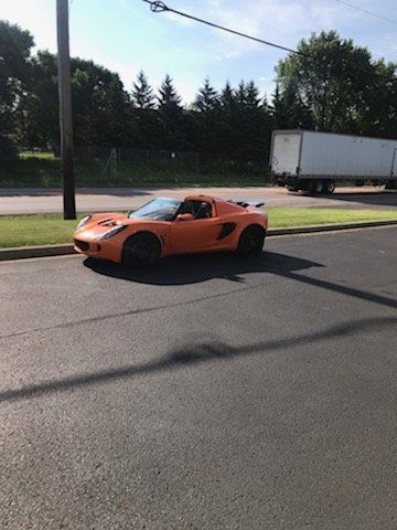 Orange sports car parked on asphalt next to a curb. Trees and a truck trailer in background.