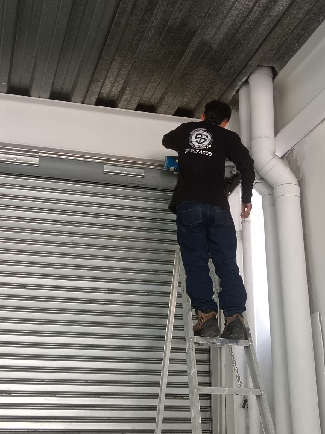 Un trabajador, subido a una escalera, inspecciona las tuberías del techo junto a una puerta metálica enrollable.