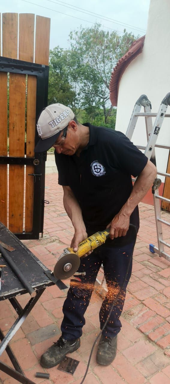 Un trabajador corta metal con una amoladora angular al aire libre; saltan chispas junto a una valla de madera.