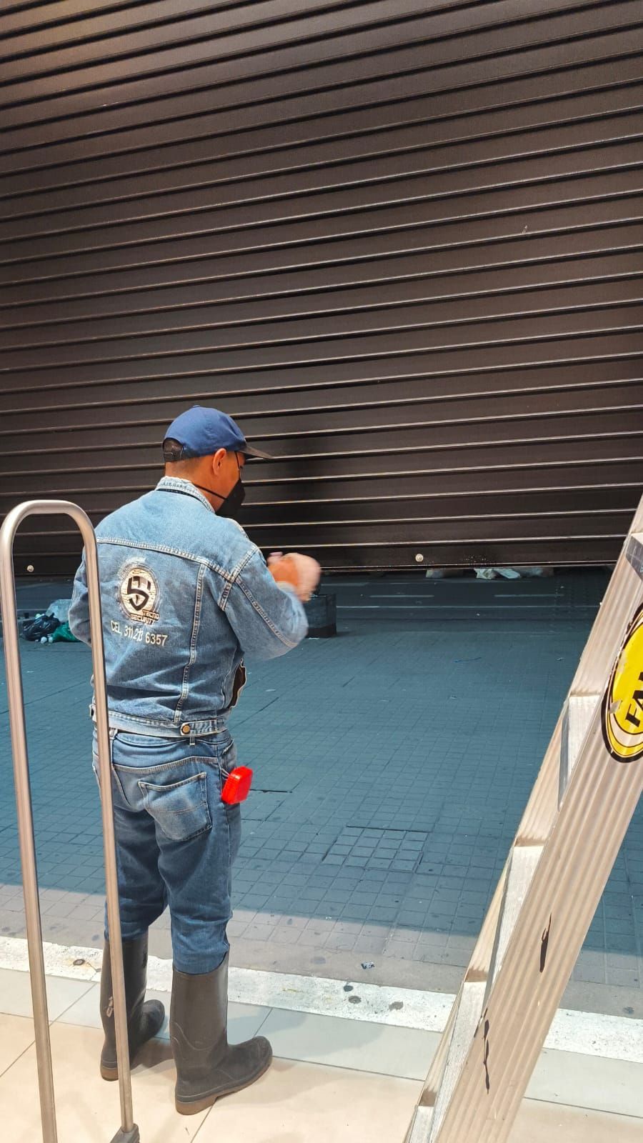 Un trabajador con uniforme azul abre una puerta enrollable de metal oscuro con una escalera cerca.