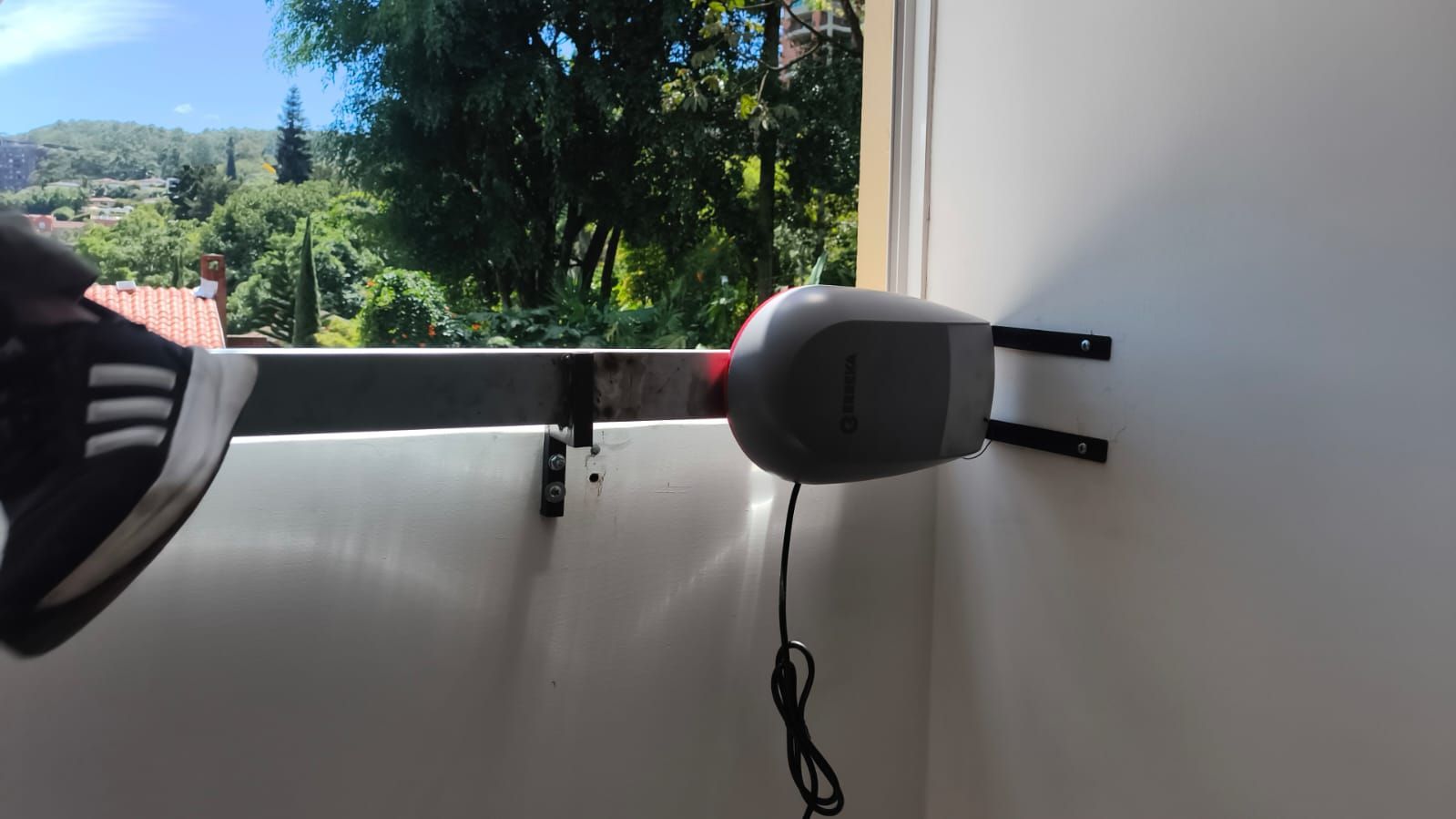 Un altavoz gris montado en la pared junto a una ventana, con un cable colgando debajo y vegetación en el exterior.