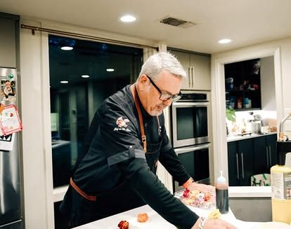 Man in a black jacket preparing food in a modern kitchen, leaning over a counter.