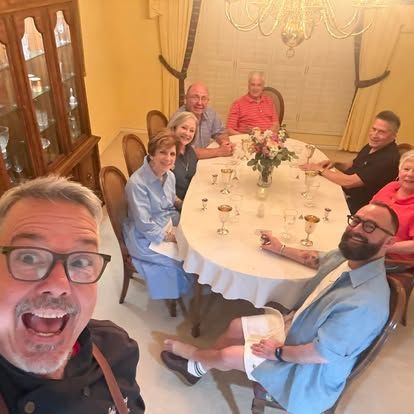 Group selfie around a dining table in a warm, elegant dining room, with smiling guests and a flower centerpiece.