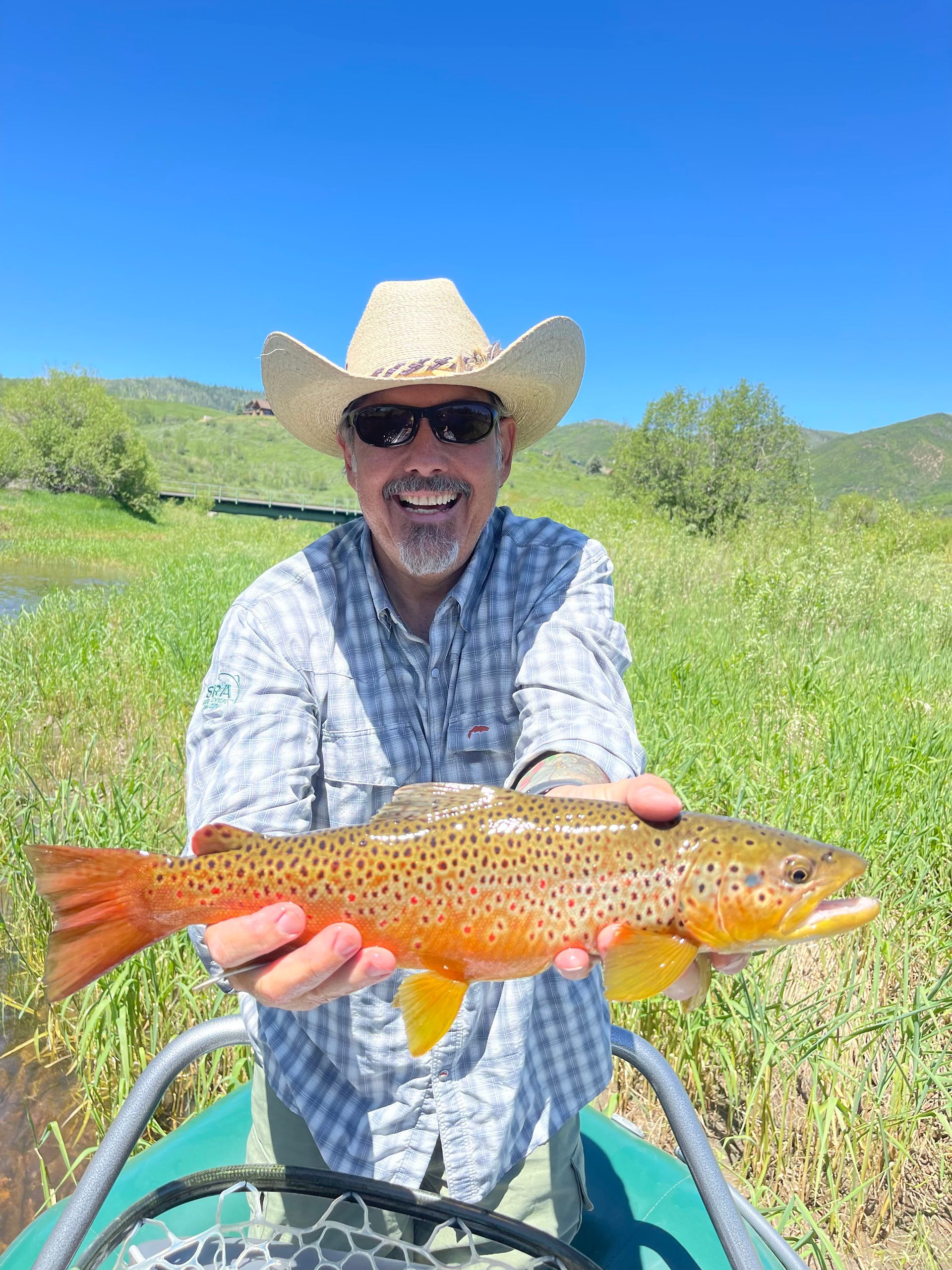 Smiling angler in straw hat holds an orange speckled trout beside a stream under a blue sky