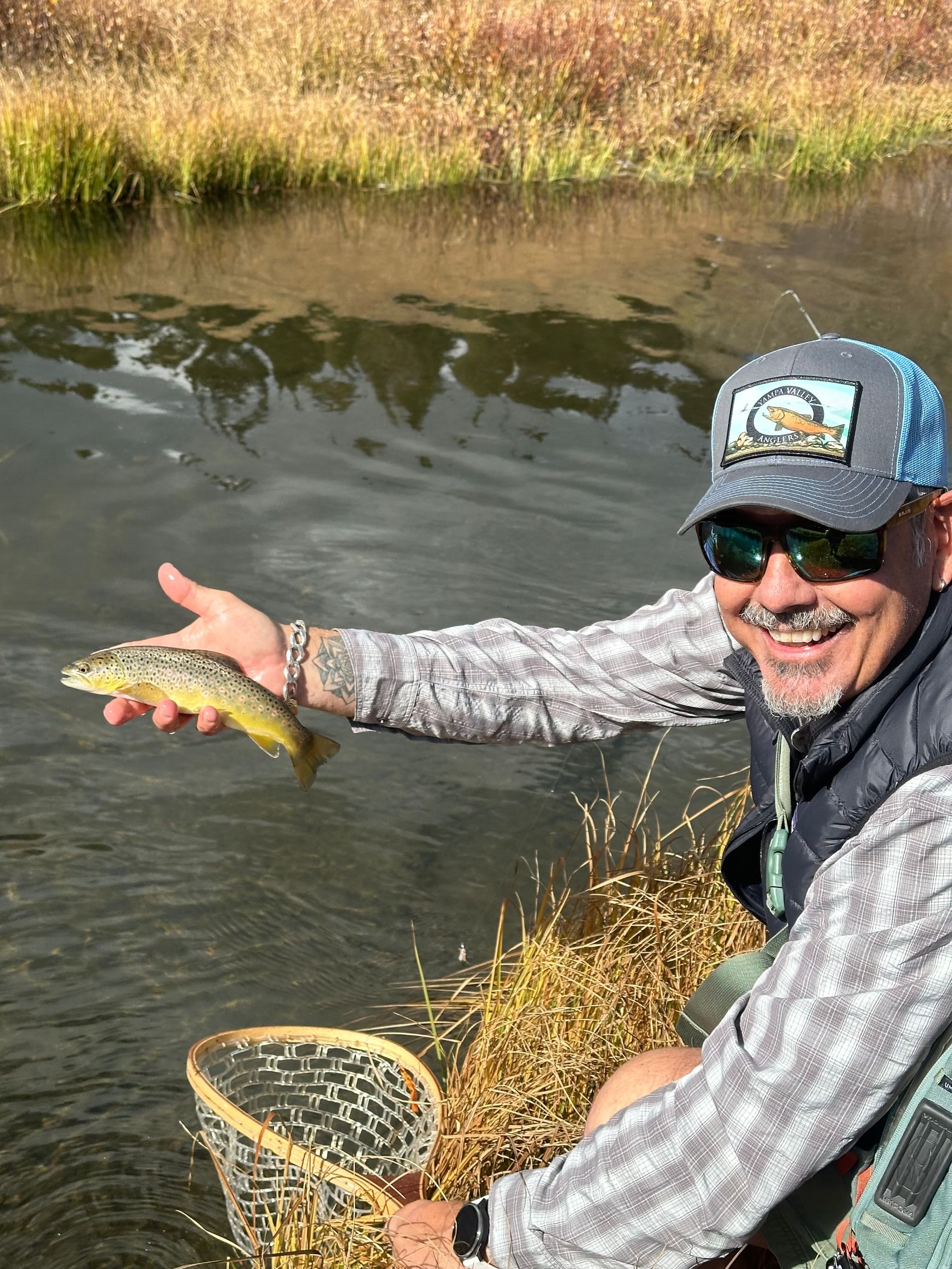Smiling angler holding a small fish beside a net in a shallow riverbank