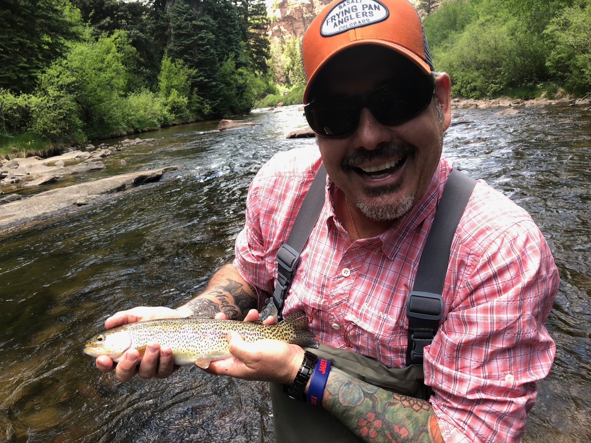 Smiling angler in orange cap holding a fish in a shallow stream, surrounded by green trees.