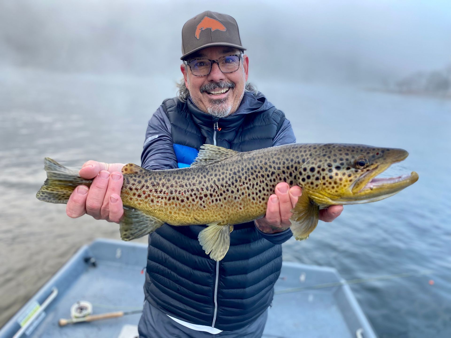 Man holding a large brown trout on a misty lake in a boat, smiling at the camera
